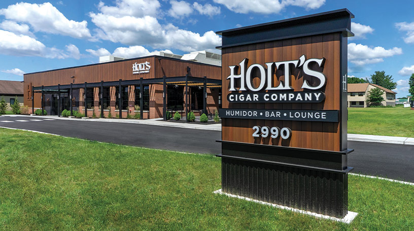 Holt's Northeast: a single-story building with brown siding, set back from the road with parking out front and a freestanding Holt's Cigar Company sign.